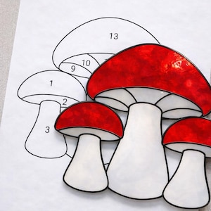 May include: A stained glass mushroom design with red caps and white stems. The mushrooms are outlined in black and sit on a white paper with numbered sections. The design is for a craft project.