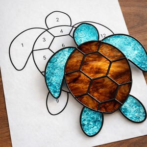 May include: A stained glass turtle with a brown shell and blue flippers rests on a numbered pattern. The turtle's shell is a mosaic of brown and amber hues, while the flippers are a vibrant blue. The pattern is a black outline on white paper.