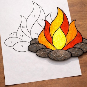 May include: A colorful stained glass campfire design. The flames are a mix of yellow, orange, and red, resting on dark gray stones. A partially completed line drawing of the same design is on a white sheet of paper.