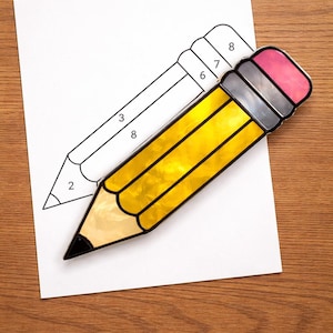Puede incluir: Un lápiz de vidriera con cuerpo amarillo, punta negra, virola gris y goma rosa descansa sobre papel blanco con un contorno de lápiz. El lápiz es un artículo decorativo.