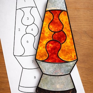 May include: A stained glass lava lamp with a vibrant orange and red design. The lamp has a black outline and a silver base and top. A paper with a line drawing of the lamp is next to it. The lamp is on a wooden surface.