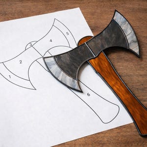 May include: A stained glass axe with a wooden handle rests on a white paper with an axe outline. The axe head is gray and black, and the handle is brown. The paper has numbers labeling the axe design.