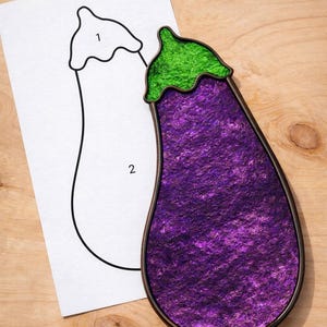 Pode incluir: Objeto decorativo em forma de berinjela, com um corpo roxo texturizado e uma parte superior verde. O objeto é delineado por uma moldura marrom escura. Ao lado, há um papel com um contorno de berinjela.