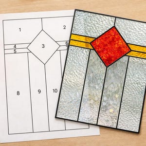 May include: A stained glass panel with a red diamond center and clear textured glass, set in a black frame. A paper pattern with numbered sections lies beside it, on a wooden surface. The panel has yellow accents.