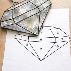 May include: A diamond-shaped stained glass ornament with a black metal frame and a chain for hanging. The glass has a clear, iridescent finish. A diagram of the diamond shape is on white paper, with numbered sections.
