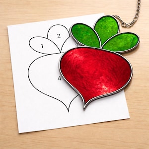 May include: A stained glass radish suncatcher with a silver-colored metal frame and chain. The radish is red with green leaves. A paper with a radish outline and numbers is next to the suncatcher.