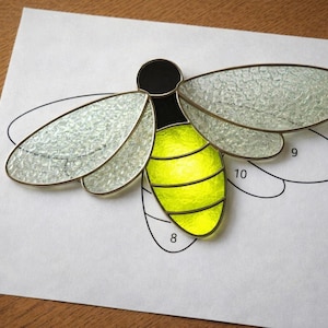 May include: A stained glass firefly with a yellow abdomen, black head, and clear wings with a textured appearance. The firefly is placed on a white paper with a black outline of the firefly's wings.