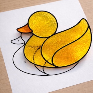 May include: A yellow stained glass duck with black outlines, resting on a white paper with a numbered outline. The duck has an orange beak and is made of textured glass. The image is on a wooden surface.