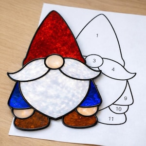 May include: A stained glass gnome with a red hat, white beard, blue jacket, and brown boots. A paper template with numbered sections is next to the gnome. The gnome is on a light wood surface.