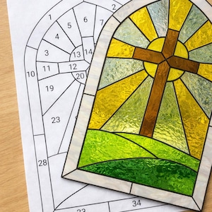 May include: A stained glass window with a cross design, featuring yellow, green, and brown colors. A paper with a numbered outline for the stained glass is next to the window. The window has a decorative chain for hanging.