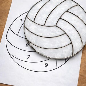 May include: A silver-toned metal coaster shaped like a volleyball, on a paper with a volleyball outline and numbers. The coaster has a white, textured interior and a black outline. The numbers 1, 7, 8, and 9 are visible.