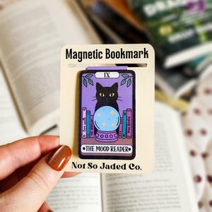 Könnte beinhalten: Magnetischer Lesezeichen mit einer schwarzen Katze und einer Kristallkugel, betitelt "The Mood Reader". Das lila-cremefarbene Lesezeichen trägt die Aufschrift "Magnetic Bookmark" und "Not So Jaded Co."