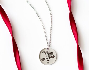 Puede incluir: Un collar de plata con un colgante redondo que presenta un delicado diseño floral. El colgante está grabado con un ramo de flores. El collar se muestra en una cinta roja.