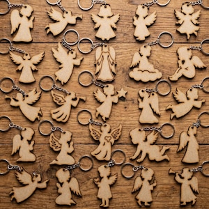 Puede incluir: Una colección de llaveros de ángeles de madera con anillas plateadas. Los ángeles están en varias poses, algunos sosteniendo estrellas o trompetas. Los llaveros están dispuestos sobre una superficie de madera.
