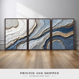 May include: Three framed abstract art prints. Each print features a layered design with shades of blue, beige, and white, resembling geological formations. The frames are dark brown. The text "PRINTED AND SHIPPED" is at the bottom.