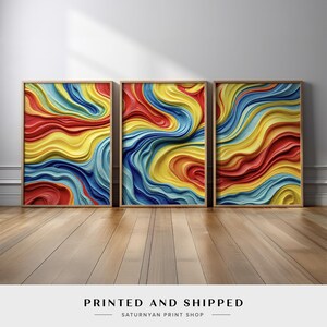 May include: Three framed abstract art prints. Each print features a colorful, textured design with flowing lines in shades of red, yellow, and blue. The frames are a light wood color, and the prints are leaning against a white wall on a wooden floor.