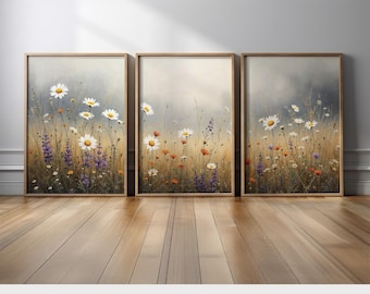 May include: Three framed art prints featuring watercolor paintings of wildflowers. The artwork showcases white daisies, purple lavender, and orange flowers in a field. The prints have a light brown wooden frame and are displayed on a wooden floor.