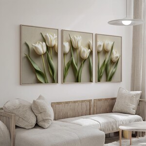 May include: Three framed wall art pieces featuring white tulip flowers with green stems and leaves. The artwork is set against a neutral background, with a light-colored frame. The art is displayed above a light-colored sofa.