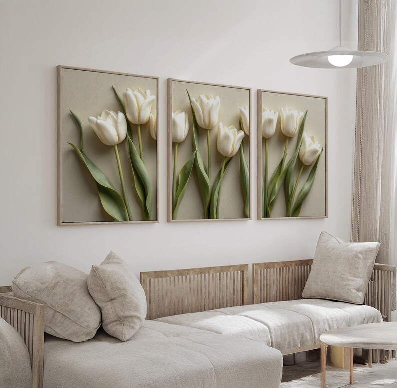 May include: Three framed wall art pieces featuring white tulip flowers with green stems and leaves. The artwork is set against a neutral background, with a light-colored frame. The art is displayed above a light-colored sofa.