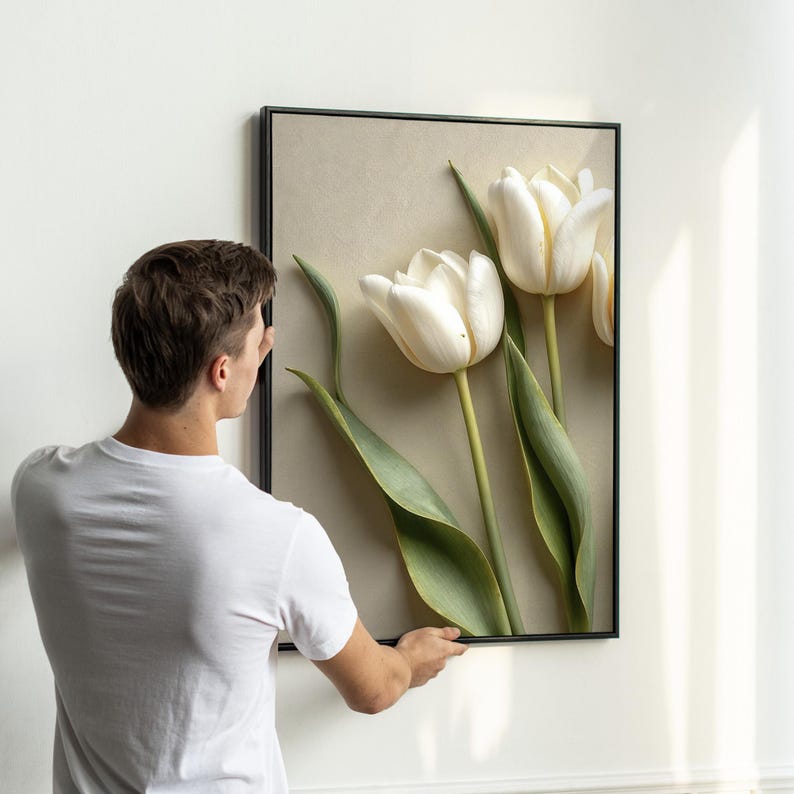 May include: A framed artwork featuring three white tulip flowers with green stems and leaves against a beige background. The artwork is in a black frame and is being held by a person wearing a white t-shirt.