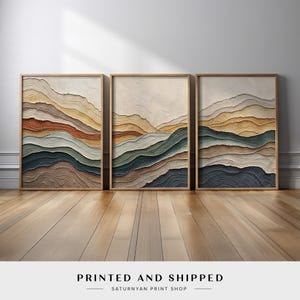 May include: Three framed abstract art prints. Each print features layered, textured designs in shades of brown, gold, green, and blue, resembling a mountain landscape. The frames are a light wood color. The text "PRINTED AND SHIPPED" is at the bottom.