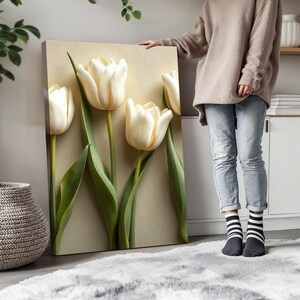 May include: A canvas art print featuring four large, creamy white tulips with green stems and leaves against a neutral background. The tulips are in full bloom, creating a sense of depth and texture. The artwork is displayed indoors.