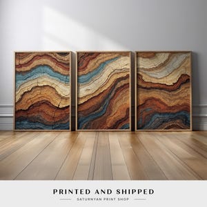 May include: Three framed art prints with abstract designs resembling tree rings. The artwork features layers of brown, orange, and blue hues. The frames are a light wood color, and the prints are displayed on a wooden floor.