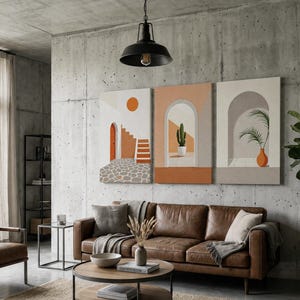 May include: A set of three rectangular wall art canvases featuring architectural scenes with arches, a staircase, a cactus in a pot, and a palm plant in an orange vase, displayed on a concrete wall above a brown leather sofa in a living room.