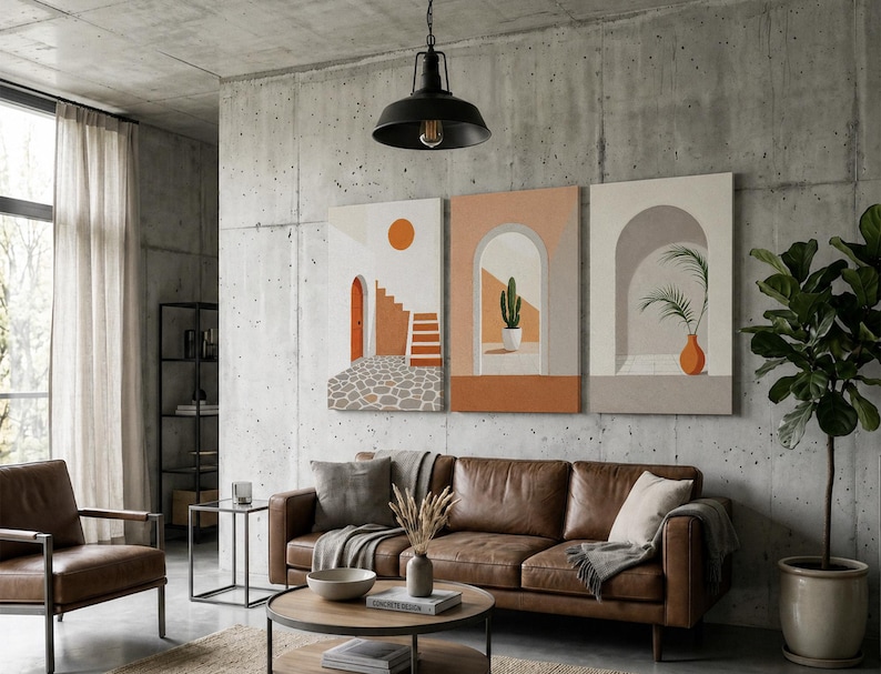 May include: A set of three rectangular wall art canvases featuring architectural scenes with arches, a staircase, a cactus in a pot, and a palm plant in an orange vase, displayed on a concrete wall above a brown leather sofa in a living room.