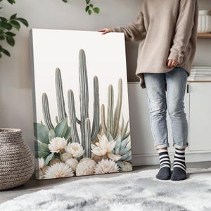 May include: A canvas print featuring a botanical illustration of cacti, succulents, and white flowers. The artwork showcases a neutral color palette with shades of green, white, and gray. The print is displayed indoors, leaning against a wall.