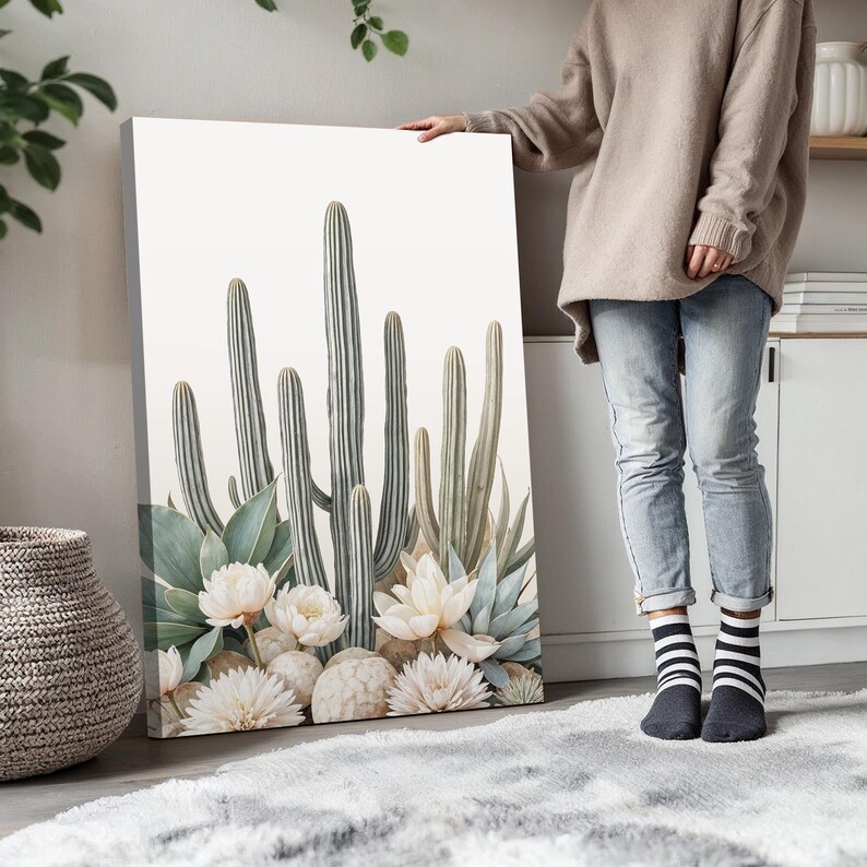May include: A canvas print featuring a botanical illustration of cacti, succulents, and white flowers. The artwork showcases a neutral color palette with shades of green, white, and gray. The print is displayed indoors, leaning against a wall.