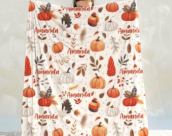 May include: A white fleece blanket featuring a repeating autumn-themed pattern of orange and white pumpkins, brown leaves, and small red berries. The name Amanda is printed in a cursive red font throughout the design, interspersed with polka dots.