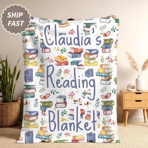 May include: A white throw blanket featuring a repeating pattern of colorful stacked books, small green leaves, and butterflies. The text "Claudia's Reading Blanket" is printed in large, dark blue script across the center of the fabric.