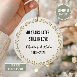 May include: A round white ceramic ornament featuring a green leaf and pink flower wreath design. The text reads: 40 YEARS LATER, STILL IN LOVE, Matias & Kate, 1986-2026. The ornament hangs from a thin gold string against a blurred background.