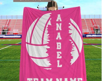 May include: A bright pink fleece blanket featuring a large white volleyball graphic split in the centre. The name ANABEL is printed vertically in white block letters between the two halves of the ball, with the text TEAM NAME at the bottom.