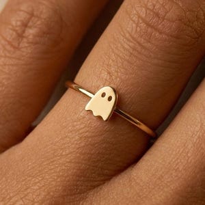 May include: A gold-colored ring featuring a small ghost charm. The ghost has two small eye holes. The ring is thin and delicate, with a simple band. The ring is shown on a person's finger.