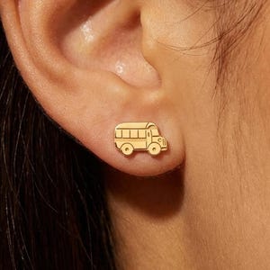 May include: Gold-colored stud earrings in the shape of a school bus. The earrings are small and detailed, with windows, wheels, and a front grill visible. The earrings are worn on an ear.