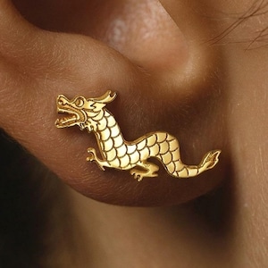 May include: A gold-colored dragon stud earring. The earring is a detailed, stylized representation of a dragon with scales, a long body, and a detailed head. The earring is worn on an ear.