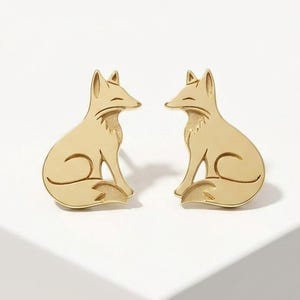 May include: A pair of gold-toned stud earrings shaped like sitting foxes. Each earring features a smooth, polished surface with engraved lines defining the fox's ears, face, and curled tail. The design is symmetrical, showing two foxes facing each other.