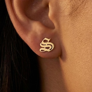 May include: A gold-colored stud earring in the shape of the letter "S" in an Old English font. The earring is small and sits on the earlobe. The background is a blurred view of a person's ear and skin.
