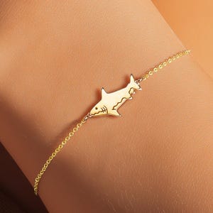 Pode incluir: Uma delicada pulseira dourada com um berloque de tubarão. O pingente de tubarão é detalhado com barbatanas e um focinho pontudo. A pulseira é usada no pulso, mostrando o design da joia. A corrente é fina e elegante.