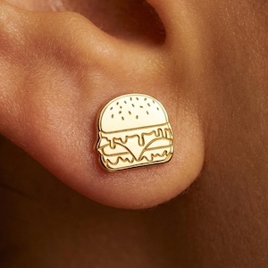 May include: A gold-colored stud earring in the shape of a hamburger, worn on an ear. The earring features a detailed outline of the burger's components, including the bun, patty, cheese, and toppings. The earring is small and delicate.