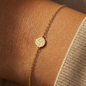 以下が含まれることがあります： 小さなバレーボールのチャームが付いた繊細なゴールドブレスレット。 チャームはバレーボールの詳細な表現であり、ブレスレットは細いチェーンです。 ブレスレットは手首に着用されており、背景はニュートラルカラーです。