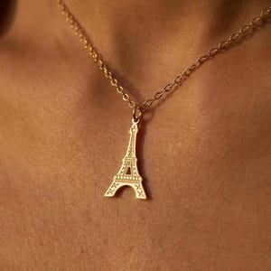 May include: A gold-toned metal necklace featuring a delicate chain and a pendant shaped like the Eiffel Tower. The pendant has a flat, cutout design with etched details representing the structure of the tower, suspended from a simple link chain.