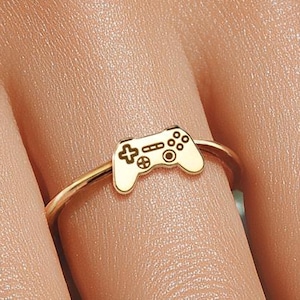 May include: A gold-colored ring featuring a small, detailed controller design. The ring's band is thin and delicate, and the controller is centered on the ring. The controller design includes buttons and a directional pad, perfect for gamers.
