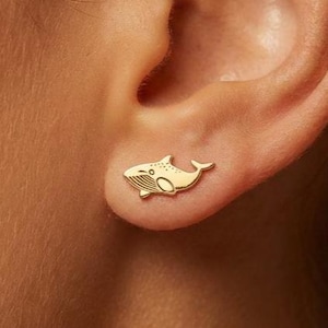 以下が含まれることがあります： 金色のクジラスタッドピアス。ピアスは小さく、詳細なクジラのデザインが特徴です。ピアスは耳に装着されており、サイズとスタイルが強調されています。シンプルでエレガントなアクセサリーです。