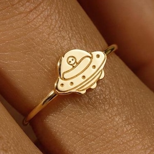 Puede incluir: Un anillo dorado con un pequeño y detallado diseño de OVNI. La banda del anillo es delgada, y el OVNI tiene un estilo de dibujos animados con una pequeña figura alienígena en su interior. El anillo se lleva en un dedo.