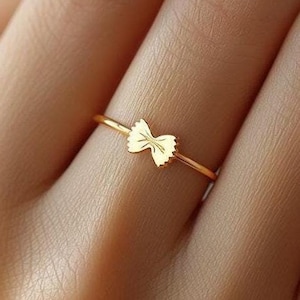 Pode incluir: Um anel de ouro com um delicado design de laço. O anel é fino e simples, o que o torna uma peça de joalheria sutil e elegante.