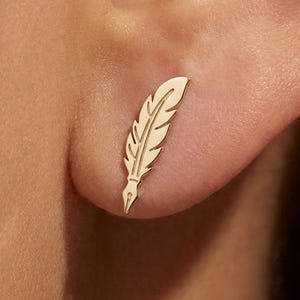 Pode incluir: Um brinco dourado em forma de pena é mostrado em uma orelha. O brinco é um brinco de pressão, com um design detalhado de uma pena. O brinco é feito de um material metálico brilhante.