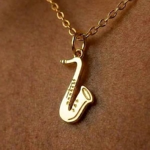 Puede incluir: Un collar de tono dorado con un pequeño colgante pulido en forma de saxofón suspendido de una delicada cadena a juego. El colgante muestra grabados detallados que representan las llaves y la campana del instrumento musical con un estilo sencillo.